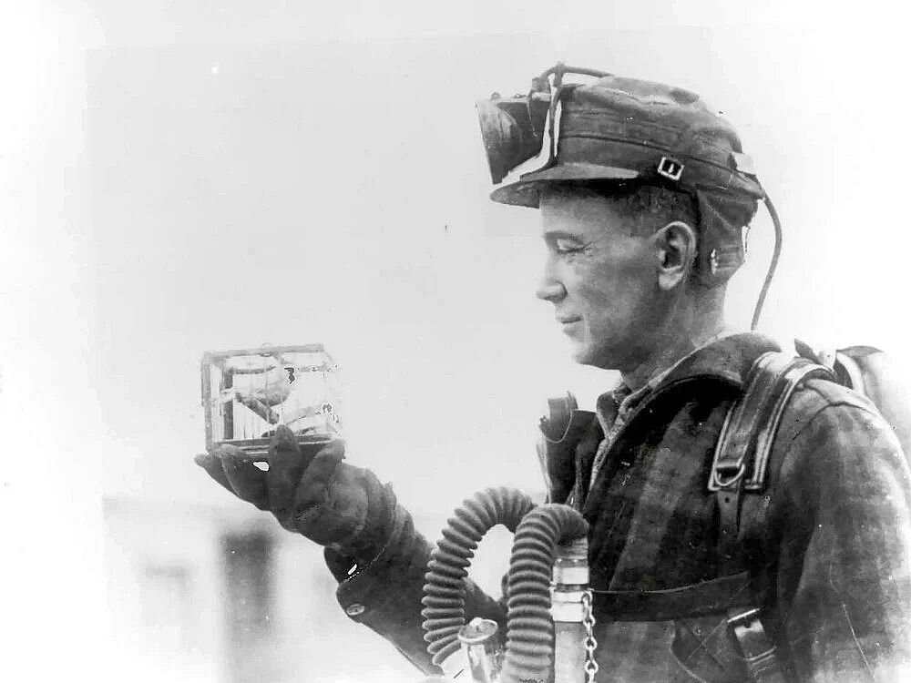 A coal miner in breathing gear holds a small caged canary in his gloved hand, reading it for danger